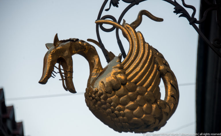 Metal dragon with open jaws, ribbed wings, and scaled body hanging from an ornate street bracket.