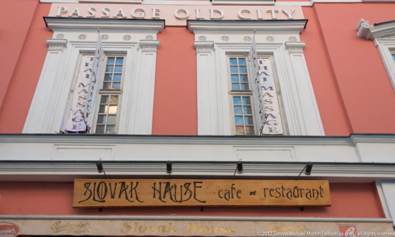 Salmon-colored Slovak House café and restaurant façade with white windows, banners, and a wooden sign.