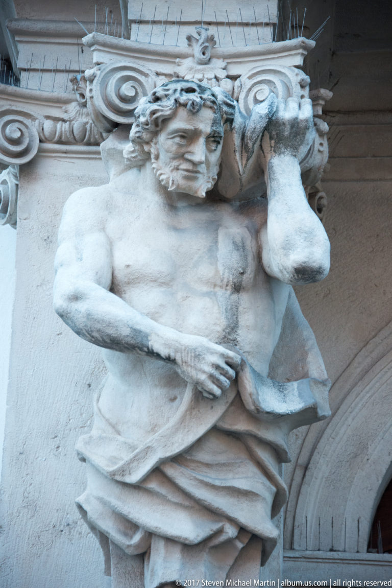 Stone male supporting figure beneath a voluted architectural capital on a decorated Bratislava façade.
