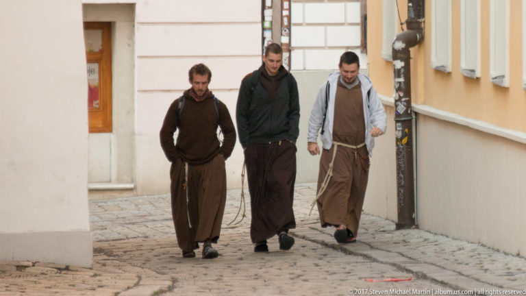 Three men in long brown rope-belted robes walking together through a narrow cobbled Bratislava street.
