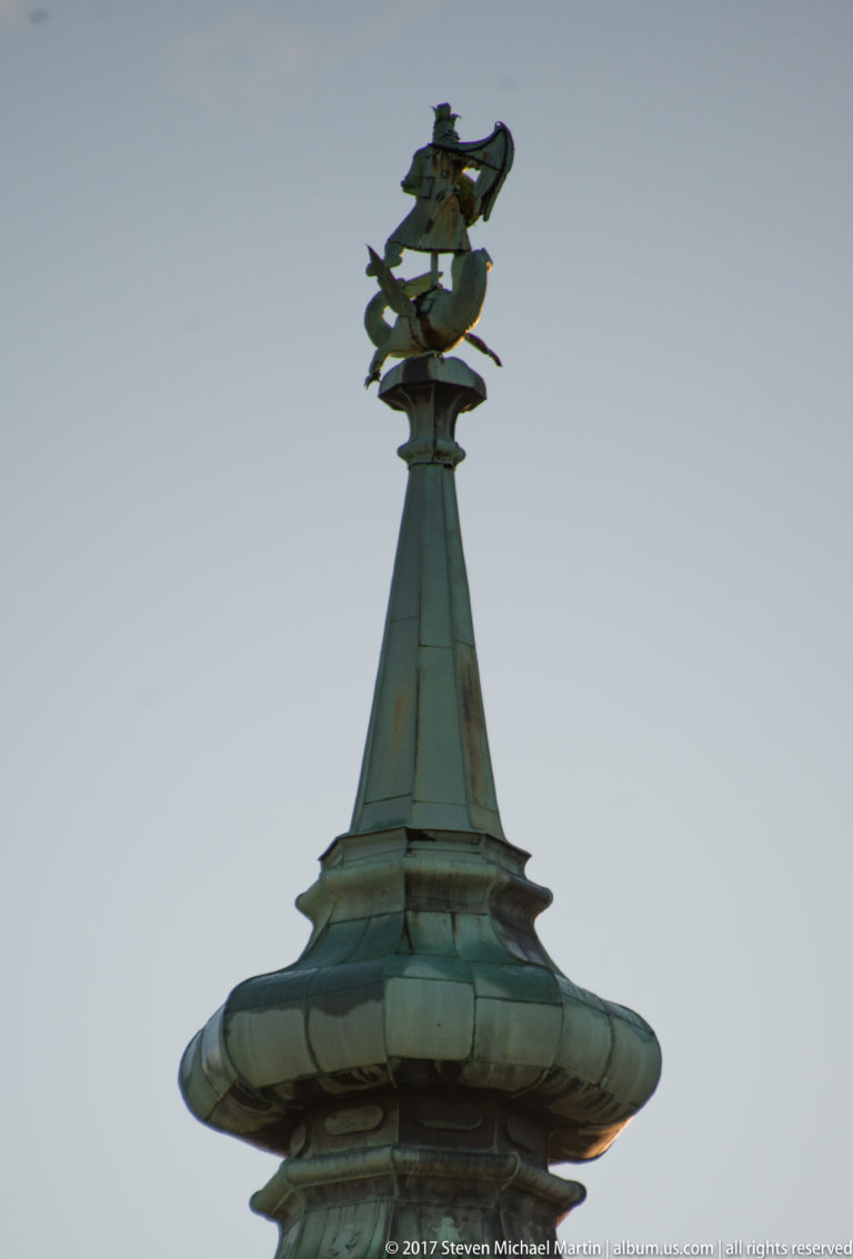 Patinated copper roof finial topped by an ornate dragon-like metal weather vane against the sky.