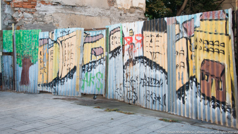 Colorful street-art mural painted across corrugated construction fencing along a paved sidewalk in Bratislava.