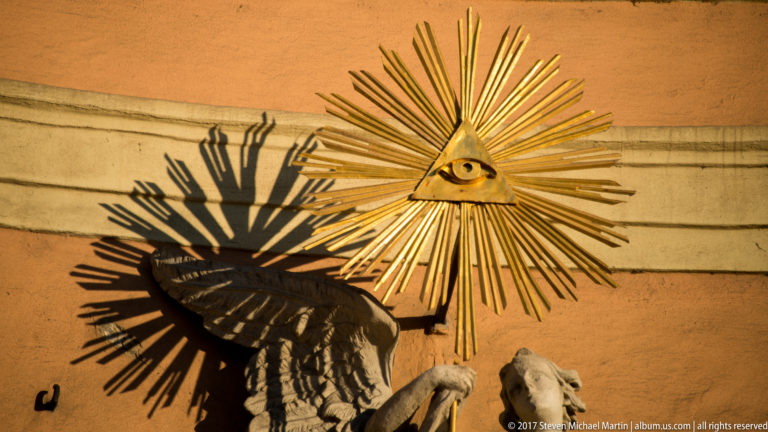 Gilded Eye of Providence in a triangular sunburst above stone angel figures on a historic Bratislava façade.