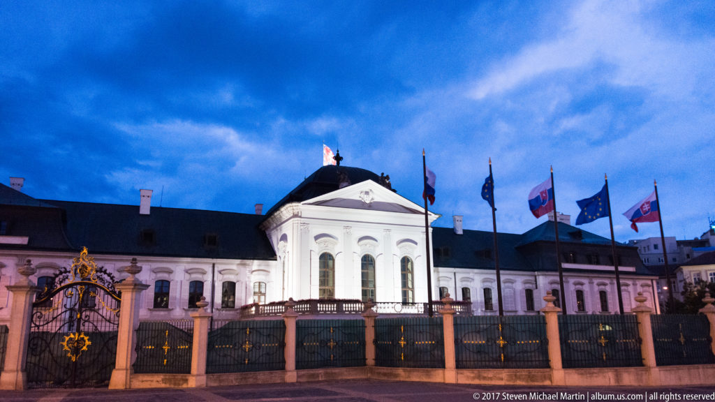 Frontal dusk view of Grassalkovich Palace behind an ornate fence and flagpoles bearing Slovak and European flags.