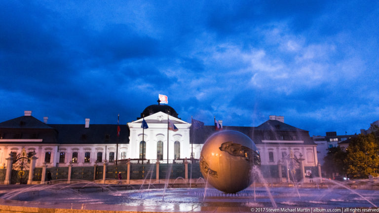 Grassalkovich Palace at dusk behind an illuminated fountain with a large spherical centerpiece in the foreground.