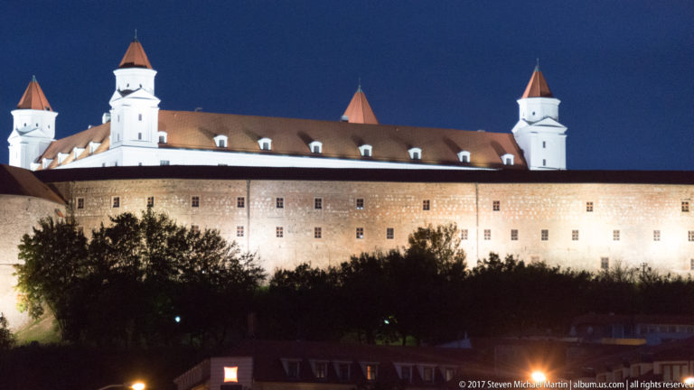 Illuminated Bratislava Castle at night, with long pale façade, red roof, and four corner towers above dark trees.