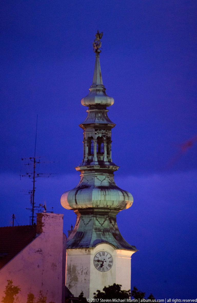 Close view of Michael's Gate tower showing its clock face, tiered green-copper roof, and crowning finial at dusk.