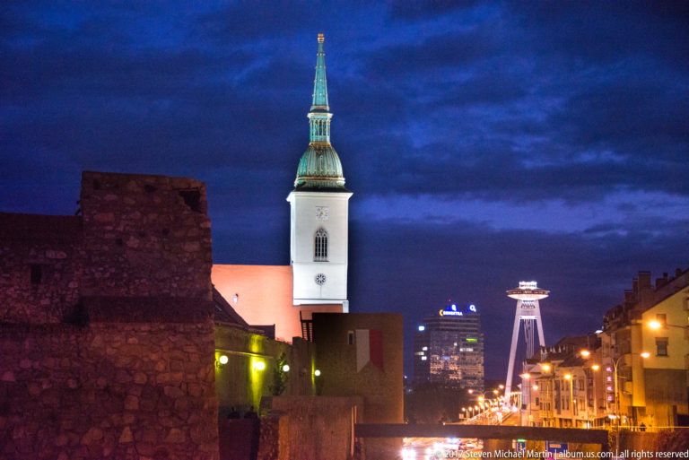 St. Martin's Cathedral tower at dusk with stone walls in front and the SNP Bridge observation tower visible in the distance.