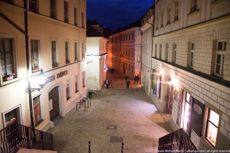 Elevated view down a cobbled old-town lane lined with pale historic façades and a few pedestrians at night.