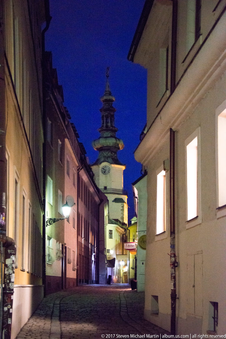 Michael's Gate tower rising at the end of a narrow cobbled lane lined with old-town buildings at night.