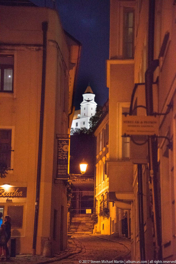 Narrow nighttime lane between historic buildings with illuminated Bratislava Castle visible high on the hill beyond.