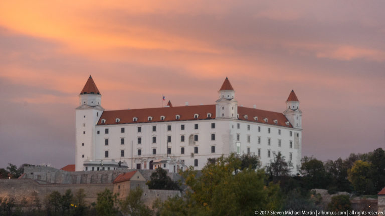 Closer sunset view of Bratislava Castle with white façade, red roof, four towers, and lower walls beneath orange-pink clouds.