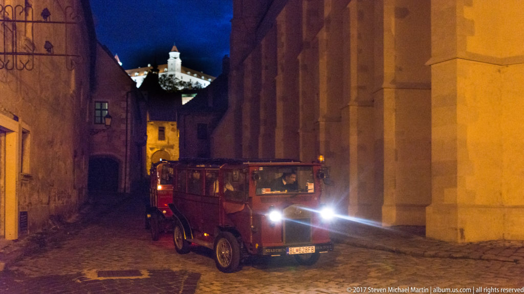 Vintage car with bright headlights in a narrow stone-lined lane, with Bratislava Castle illuminated on the hill above.