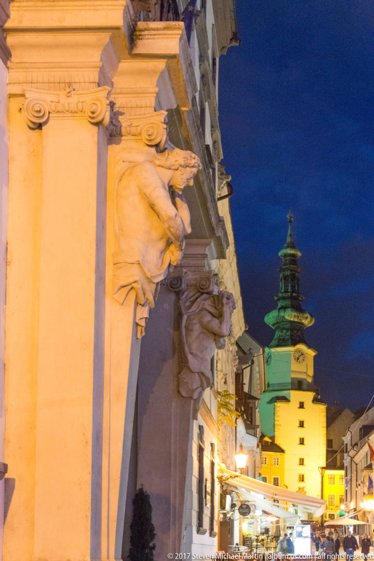 Ornate illuminated façade at left with the green-roofed Michael's Gate tower rising beyond a narrow Bratislava street at night.