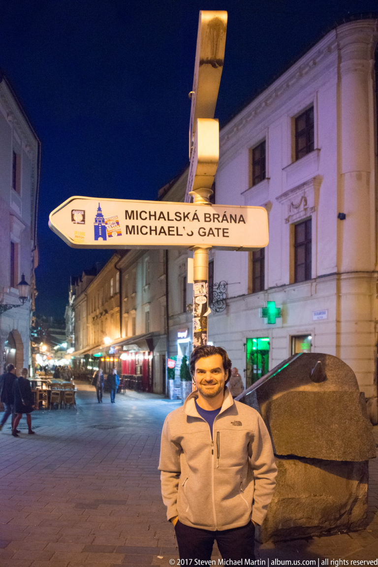 Directional sign reading “Michalská brána / Michael's Gate” above a nighttime pedestrian street in central Bratislava.