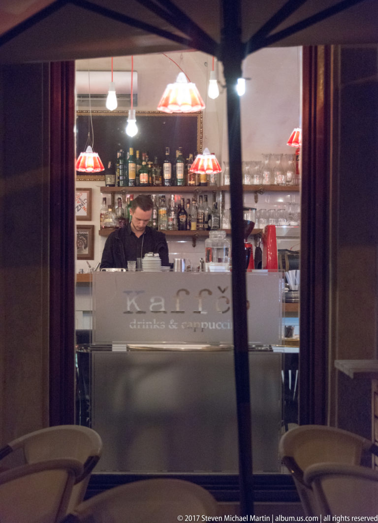 Barista or bartender behind a small counter seen through a reflective window, with hanging lamps and shelves of bottles.