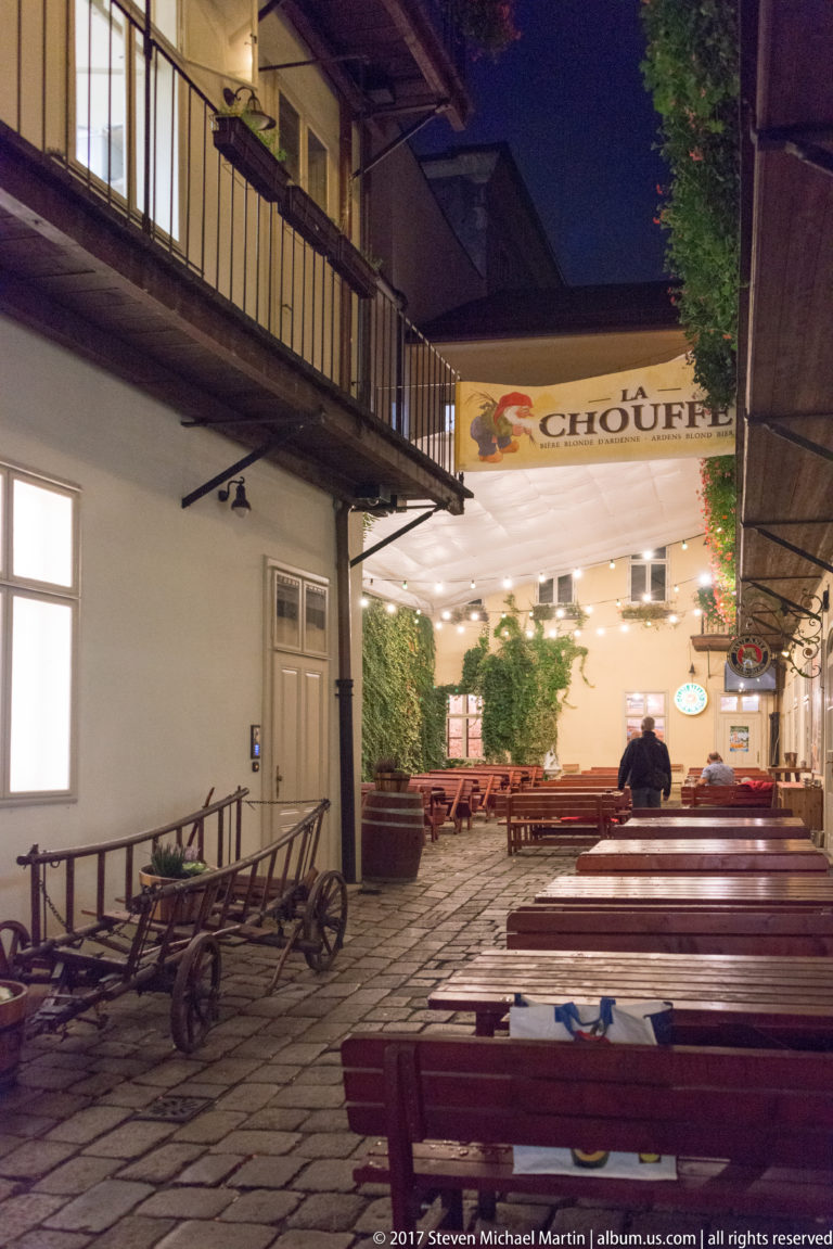 Nighttime beer garden with long wooden tables, balcony galleries, a La Chouffe sign, and an old cart along the wall.