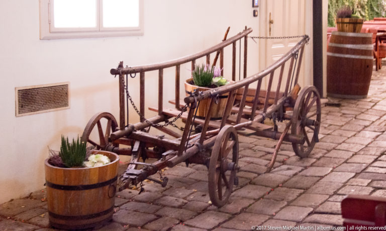 Old wooden cart with spoked wheels on cobblestones beside white walls and barrel planters in a Bratislava courtyard.