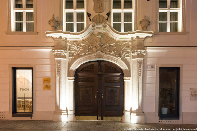 Illuminated Baroque doorway with carved scrollwork and tall dark wooden doors on a pale historic Bratislava façade.