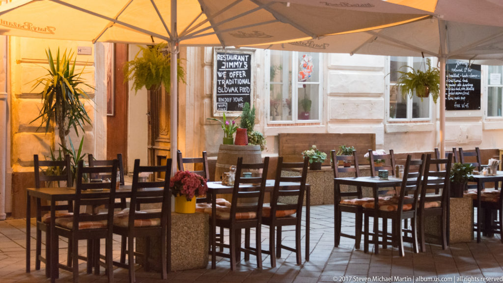 Empty outdoor restaurant terrace with wooden tables, chairs, umbrellas, menu boards, and potted plants in a Bratislava courtyard.