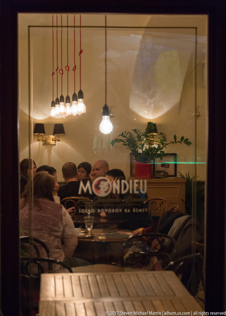 Diners inside Mondieu café seen through a large street window with pendant lights and reflections at night.