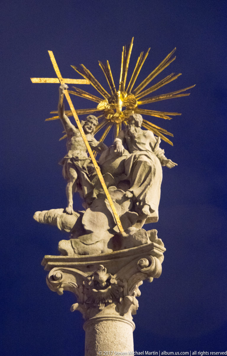 Baroque stone figures beneath a burst of gilded rays atop a devotional column, viewed from below at night.