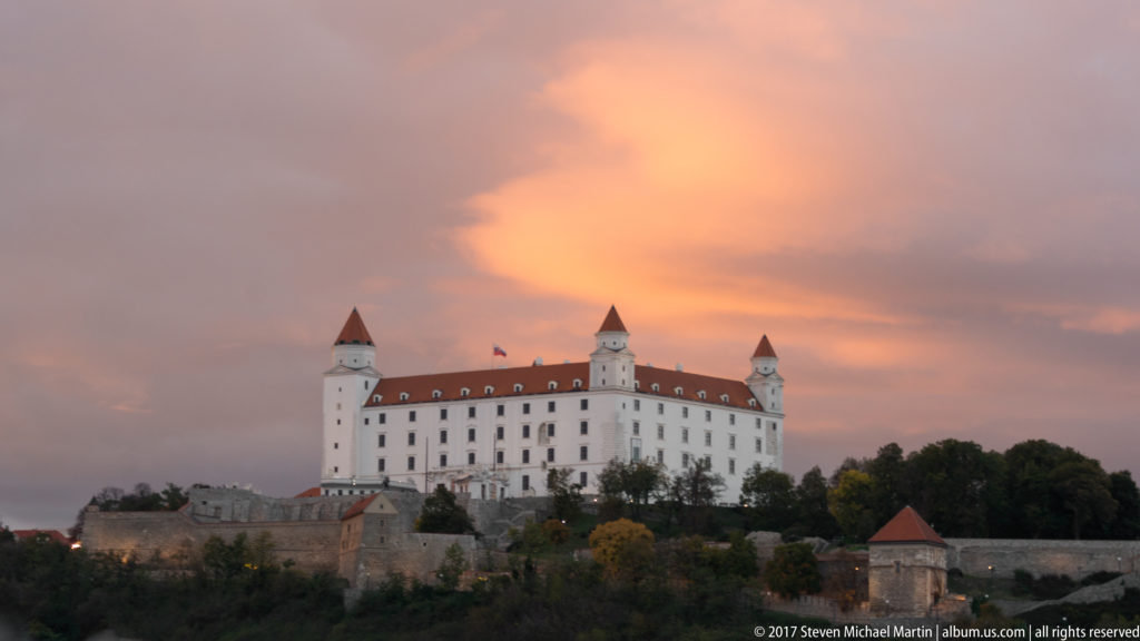 Bratislava Castle on the hill at sunset, with white walls, red roof, four corner towers, trees, and lower fortifications.