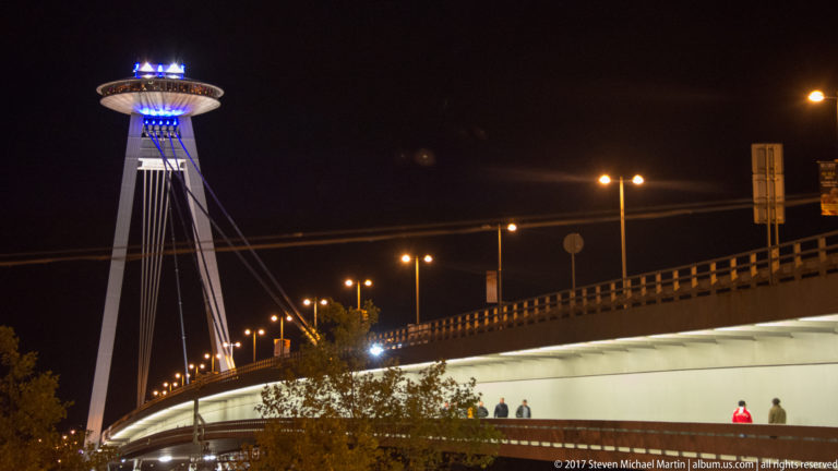 SNP Bridge at night with blue-lit UFO observation deck, angled cables, roadway, and pedestrians on the lower walkway.