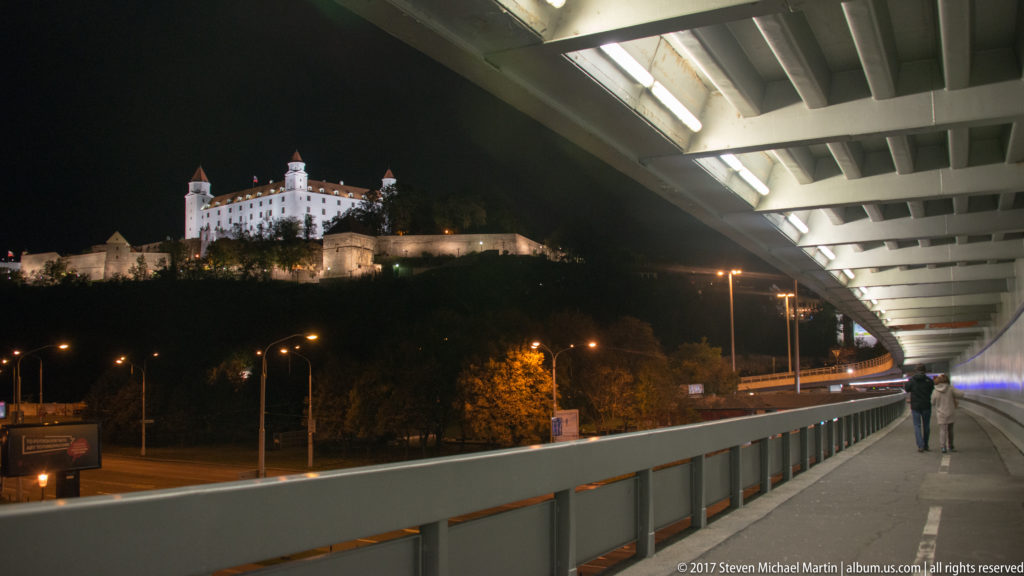 Illuminated Bratislava Castle seen from a covered SNP Bridge pedestrian approach at night, with bridge structure framing the view.