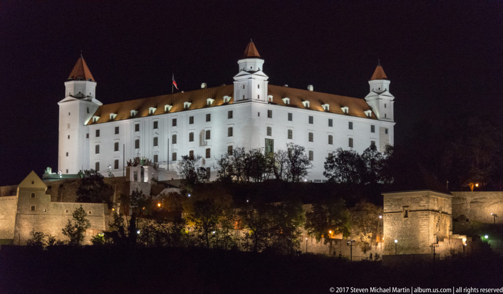 Close nighttime view of illuminated Bratislava Castle with its four corner towers above darker defensive walls and trees.