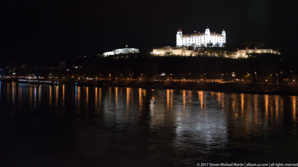 Illuminated Bratislava Castle on its hill above the Danube at night, with embankment lights reflected in the water.