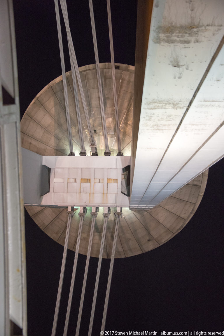 Upward view beneath the SNP Bridge showing the concrete deck, cable supports, and saucer-shaped pylon structure.