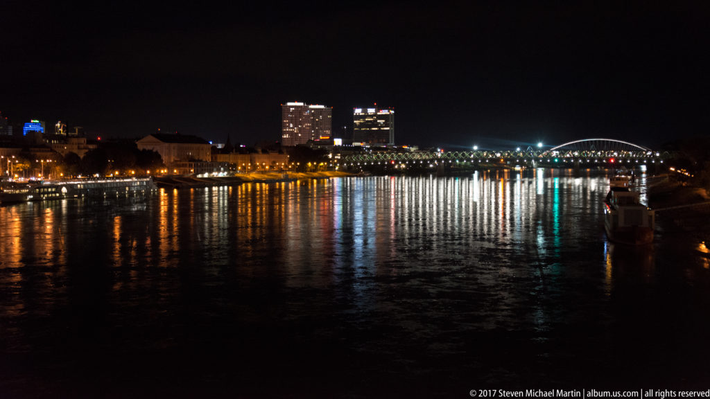 Night view across the Danube toward Bratislava’s lit riverfront and bridge, with long reflections on the water.