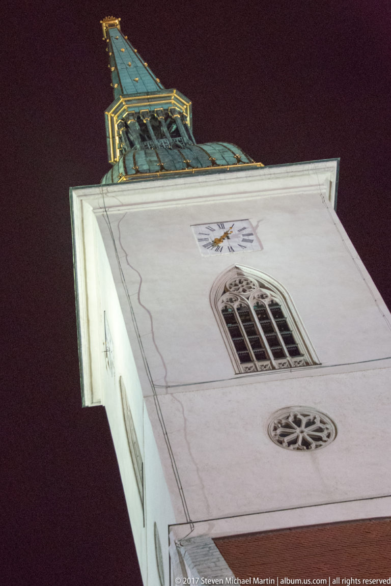 Low-angle nighttime view of a white church tower with round window, Gothic opening, heraldic emblem, and copper-green roof.