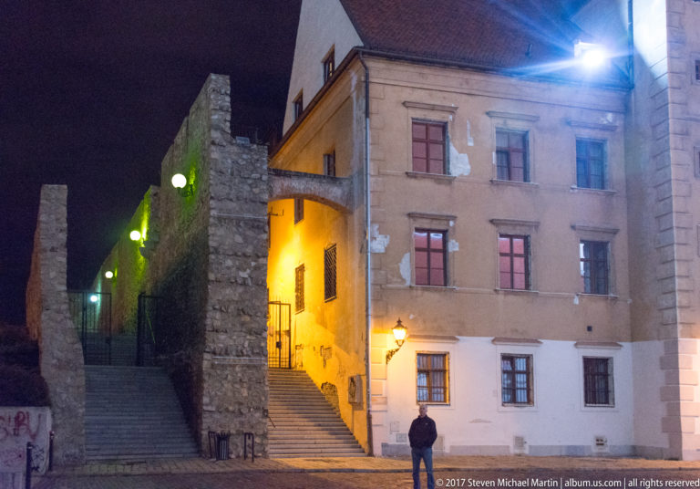 Stone steps climbing between old masonry walls and a weathered building, lit by green and amber lamps at night.
