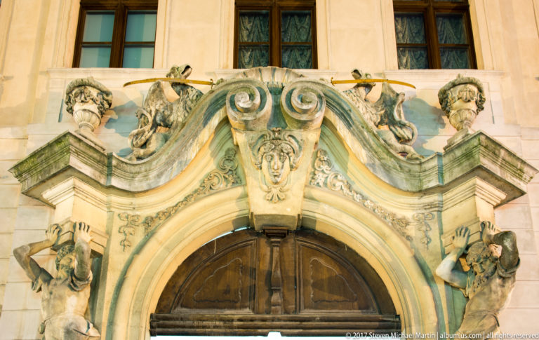 Ornate Baroque stone portal with scrolls, masks, small figures, and a dark wooden doorway below.