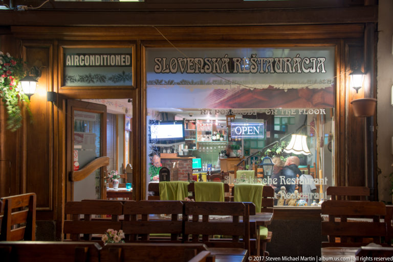 Slovak restaurant storefront at night with wooden terrace seating and a warmly lit dining room visible through glass.