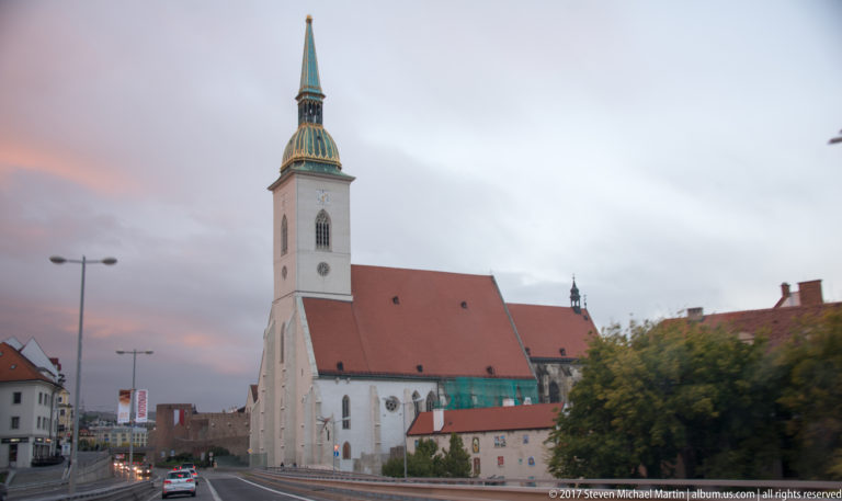 St. Martin's Cathedral with white tower, green spire, red roof, roadside traffic, and old-town walls under an evening sky.