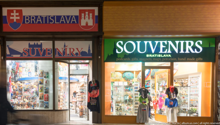 Two brightly lit Bratislava souvenir shops at night with postcards, clothing, bags, and tourist merchandise in the windows.