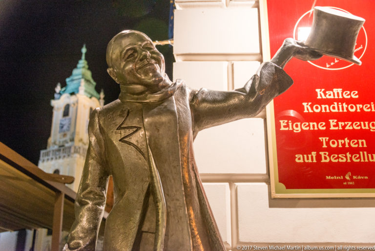 Bronze Schöne Náci figure smiling and raising his top hat on a Bratislava old-town street at night.