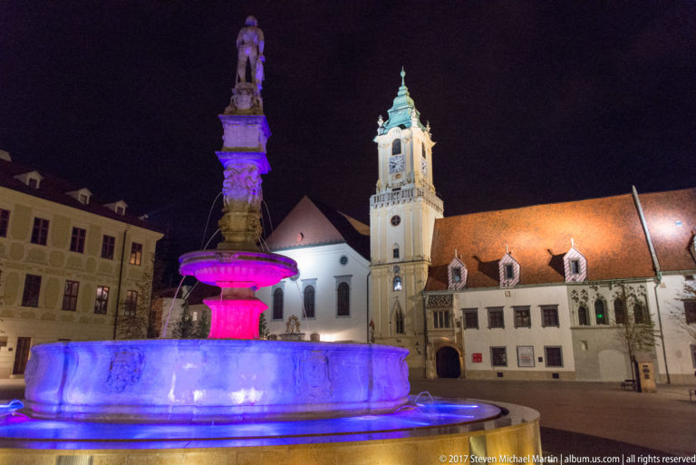 Colorfully illuminated Maximilian Fountain in Bratislava Main Square with the Old Town Hall tower rising behind.