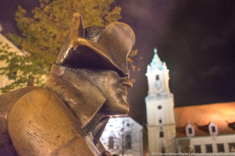 Bronze figure in a bicorne-style hat seen close-up at night with an illuminated historic tower blurred behind.
