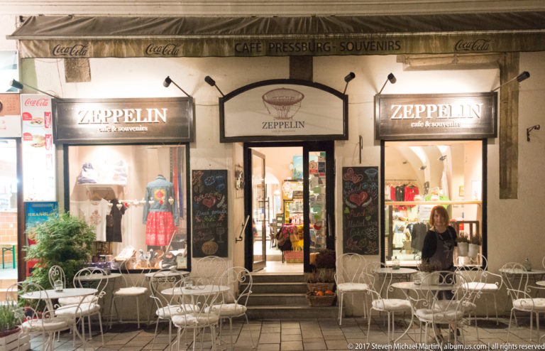 Zeppelin café and souvenir storefront at night with white outdoor tables, chairs, chalkboards, and lit display windows.