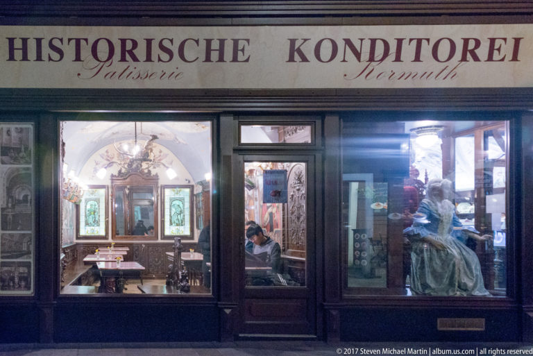Dark wood konditorei storefront at night with illuminated display windows and a person working inside.
