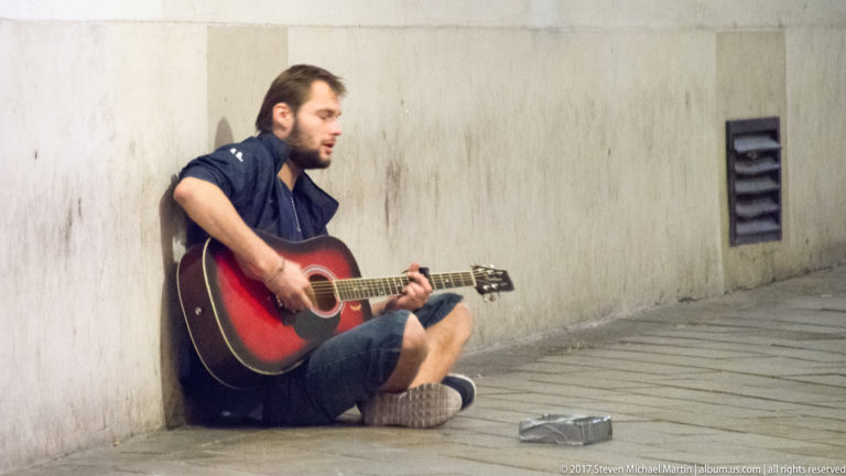 Seated street musician playing a red-and-black acoustic guitar beside a pale stone wall in Bratislava.