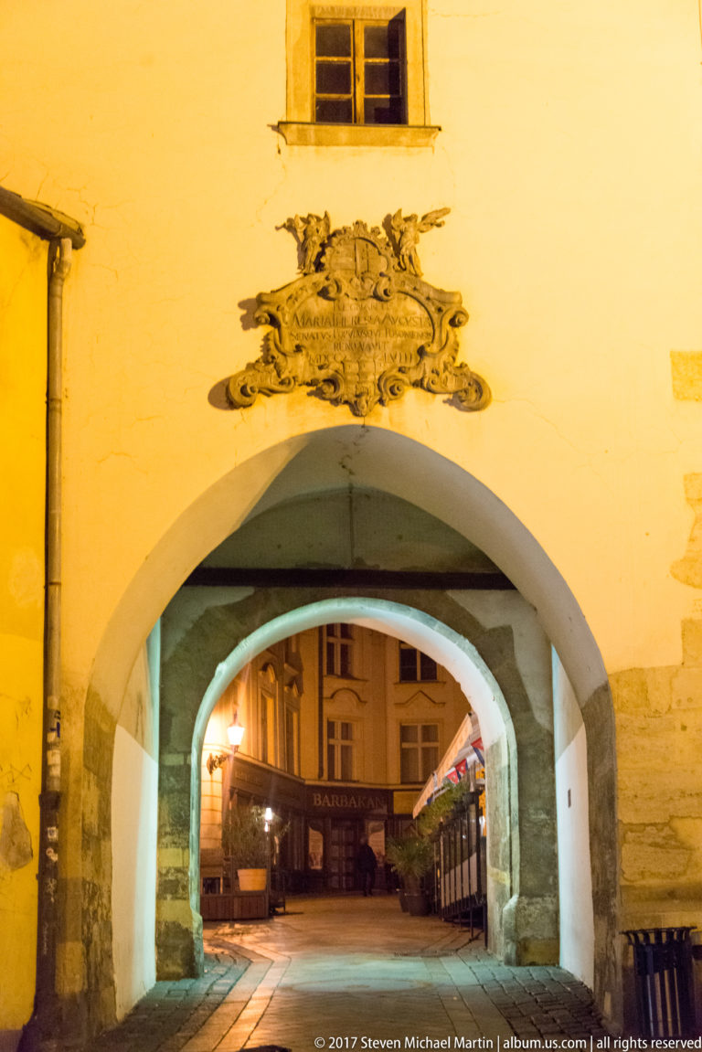 Arched passage through a yellow historic building with a heraldic relief above and a lit street beyond.
