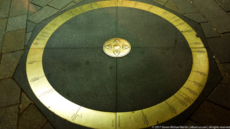 Circular brass orientation marker with a compass-like center embedded in dark stone paving.