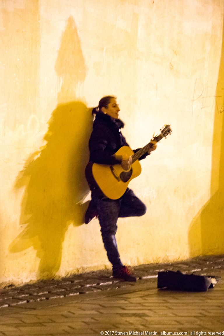 Street musician with acoustic guitar moving beside a yellow wall, casting a large dramatic shadow at night.