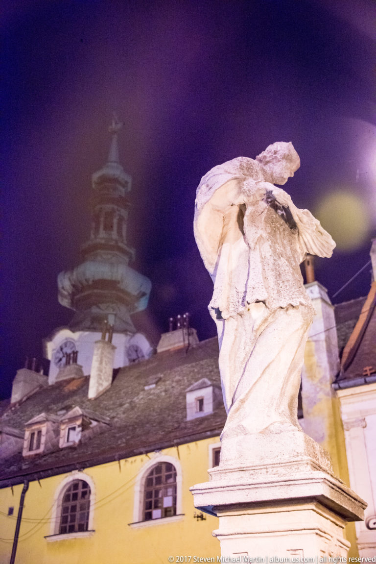 Weathered stone statue on a pedestal at night with a historic tower and roofline behind it.