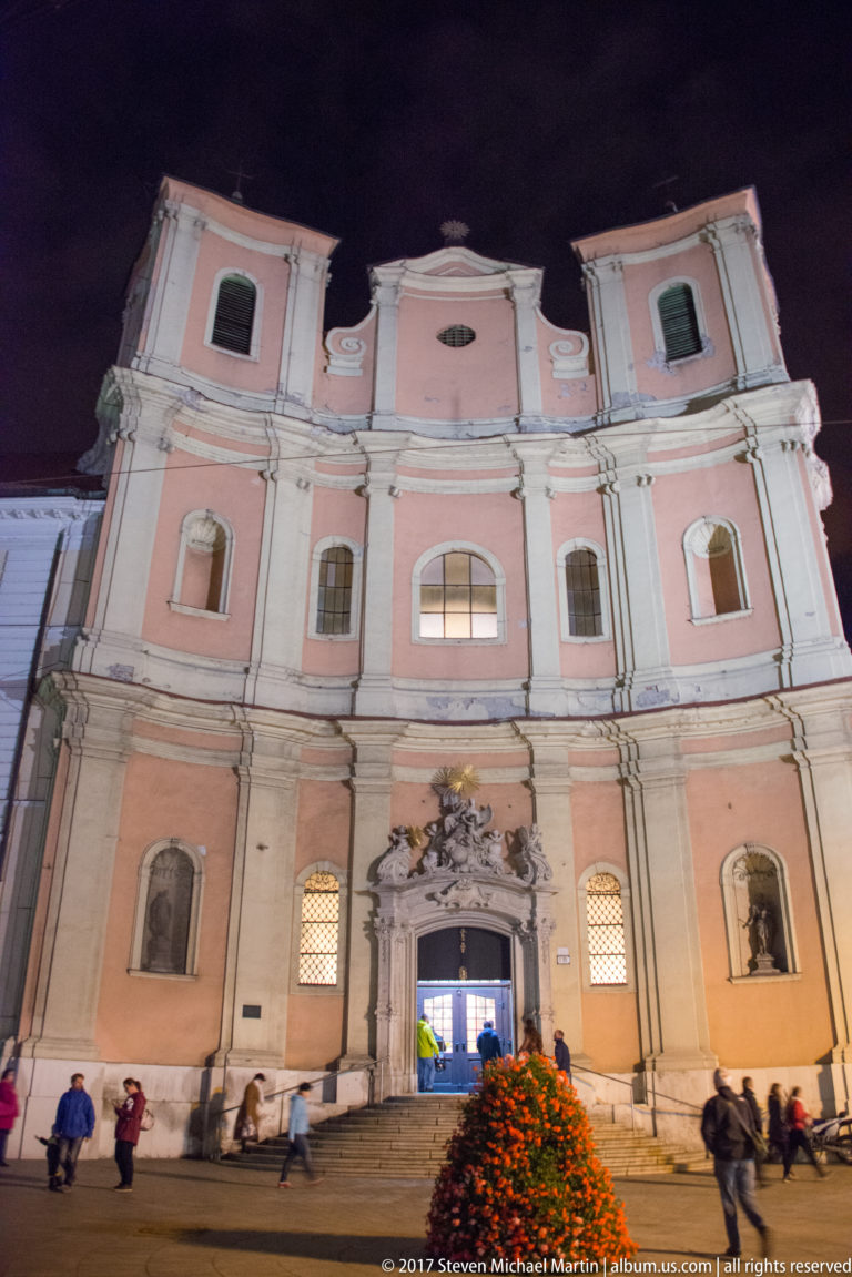 Illuminated pink-and-white Baroque church façade viewed from a small square at night with pedestrians below.